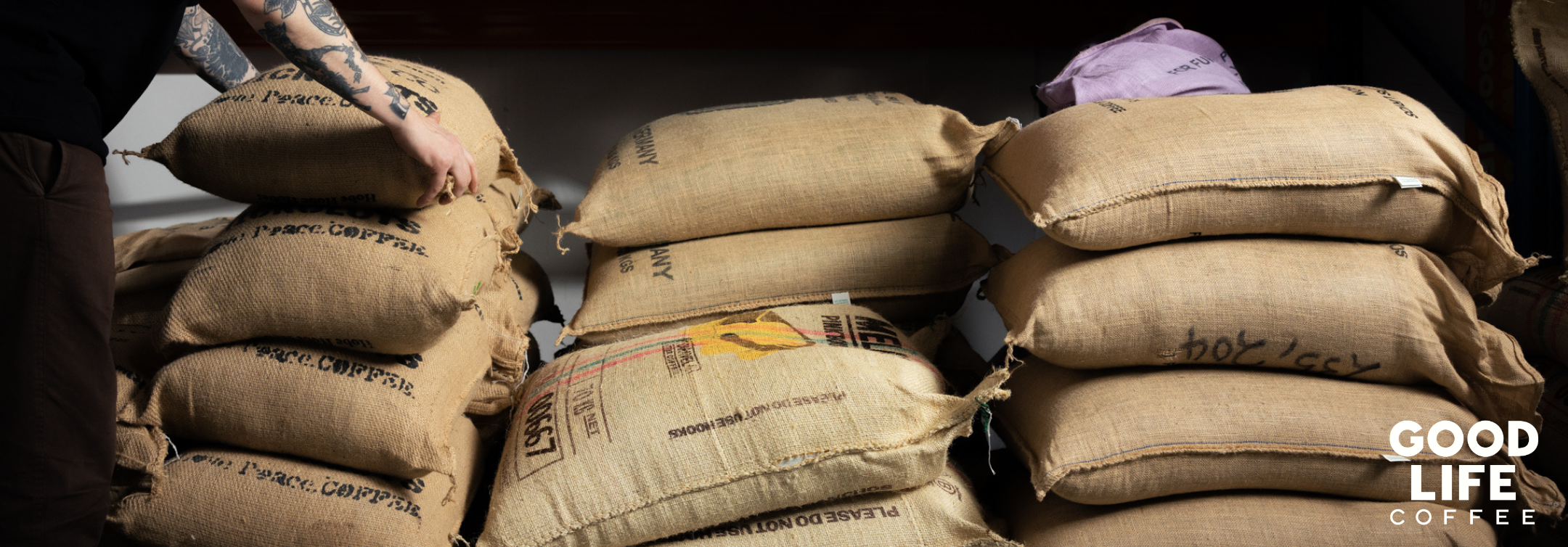 Useita kahvisäkkejä pinossa, vasemmalla henkilö siirää säkkiä, oikealla alhaalla Good Life Coffee-logo